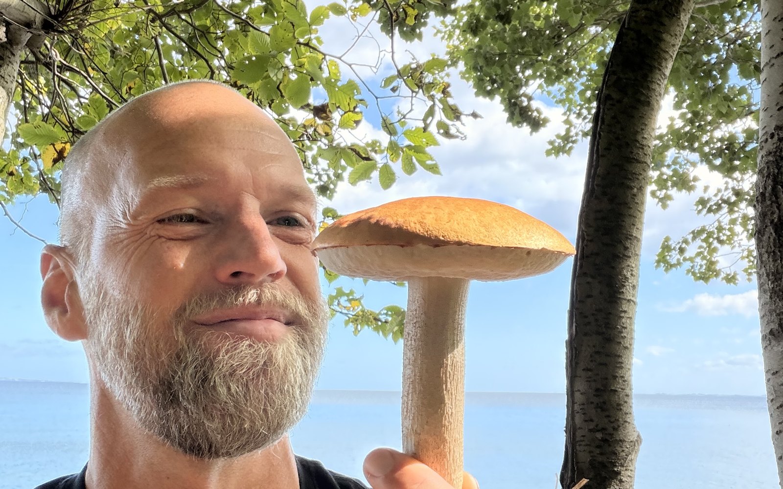 Asger i skovbrynet med en stor rørhat i hånden, havet bag træerne